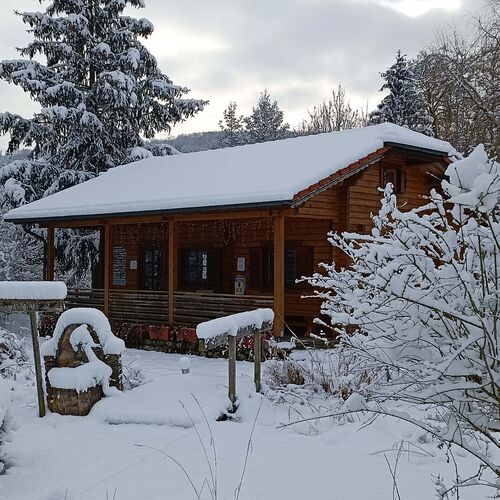 "Cozy single-story timber house with a red-tiled pitched roof, blanketed in snow, creating a picturesque winter scene. The house features a spacious covered porch supported by timber posts, adorned with delicate string lights for a warm, festive touch. Constructed from natural wood, the house blends beautifully with the snowy landscape. Surrounding the house are snow-covered plants, trees, and rustic wooden decorations, enhancing its charm and natural appeal. Supplied by Bertsch Holzbau and skillfully designed and installed by Forever Timber, this timber house offers a serene and inviting retreat in a winter wonderland."
