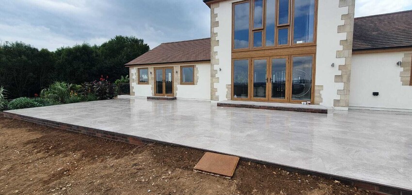Stunning patio installation featuring polished stone tiles that seamlessly extend from the elegant cream and stone-clad home. The large outdoor space is framed by lush greenery, offering a perfect setting for relaxation or entertaining with panoramic views of the surrounding landscape.