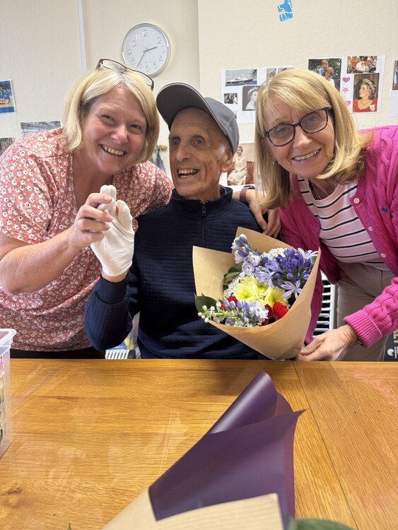 A guest smiles with two team members and a bouquet of flowers at Your Care Dementia Day Club in Hanham, Bristol, where familiar faces and personalised support help people feel welcome.