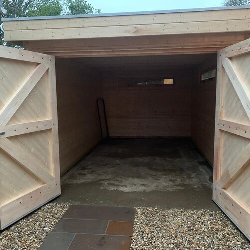 "Custom-built timber garage with a flat roof and wide double doors featuring a natural wood finish and a traditional cross-brace design. The interior is spacious and fully enclosed with high-quality wood paneling, providing a warm and durable structure. The garage is set on a solid concrete base with a gravel and paved entrance for added practicality and visual appeal. Supplied by Lugarde and meticulously designed and installed by Forever Timber, this garage offers a perfect balance of rustic charm and modern functionality."