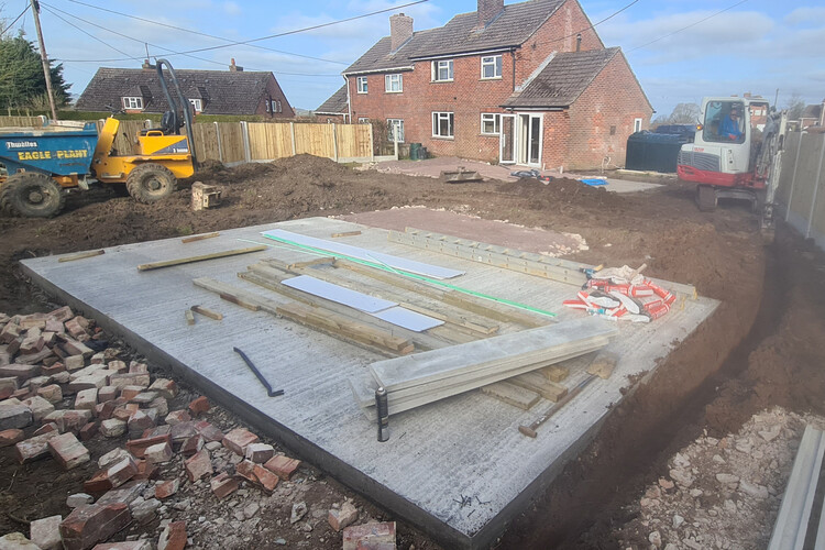 Construction site showing a newly poured concrete foundation, prepared for the installation of a building. The site is surrounded by construction materials, including timber planks, insulation boards, and tools. Heavy machinery, such as a dumper truck and an excavator, are present, emphasizing active progress on the project. The site is adjacent to a brick residential property with a neatly fenced backyard, indicating a suburban setting. This foundation marks the initial stage of creating a functional and durable structure