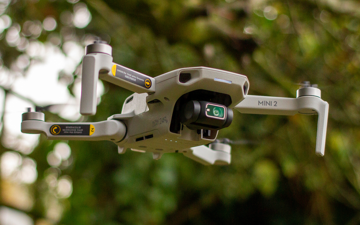 Close-up image of a DJI Mini 2 drone in flight, featuring a compact and lightweight design with a sleek gray body weighing under 249 grams. The drone is equipped with a high-quality 4K camera mounted on a 3-axis gimbal for stable and sharp aerial footage. Visible arm labels guide setup and folding instructions, emphasizing its portability and user-friendly design. The background features a blurred natural setting, highlighting the drone's versatility for outdoor photography and videography