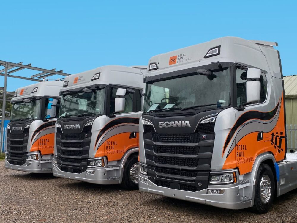 Fleet of several Scania trucks with matching branded silver and orange vinyl liveries.