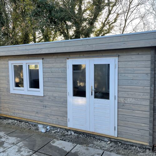 A spacious and functional wooden garden building designed and installed by Forever Timber, supplied by Lugarde. It features a light grey exterior with white accents, including double glass-panel doors and a double-pane window for enhanced natural light and ventilation. The flat roof and minimalist design make it a perfect choice for a home office, workshop, or versatile outdoor living space.