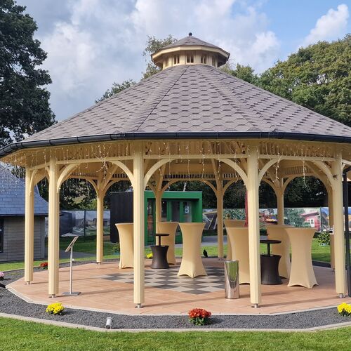 Elegant octagonal timber gazebo with a decorative cupola roof, constructed from cream-colored timber. The structure features intricate archways and open sides, creating a spacious and airy design ideal for outdoor gatherings or events. The roof is covered with durable shingles, adding a touch of sophistication. The interior includes a wooden deck with checkerboard-style detailing and is furnished with tall cocktail tables, dressed in neutral-colored linens, enhancing its versatility for various occasions. Surrounded by a manicured lawn and vibrant flower beds, this gazebo was expertly designed and installed by Forever Timber, combining timeless beauty with superior craftsmanship.