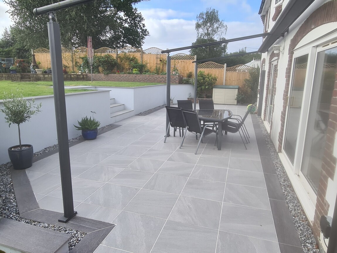 Stylish outdoor patio with large modern grey tiles, complemented by sleek black garden furniture. The multi-tiered design features white retaining walls and neatly landscaped greenery, creating a functional yet elegant space for relaxation and entertaining.