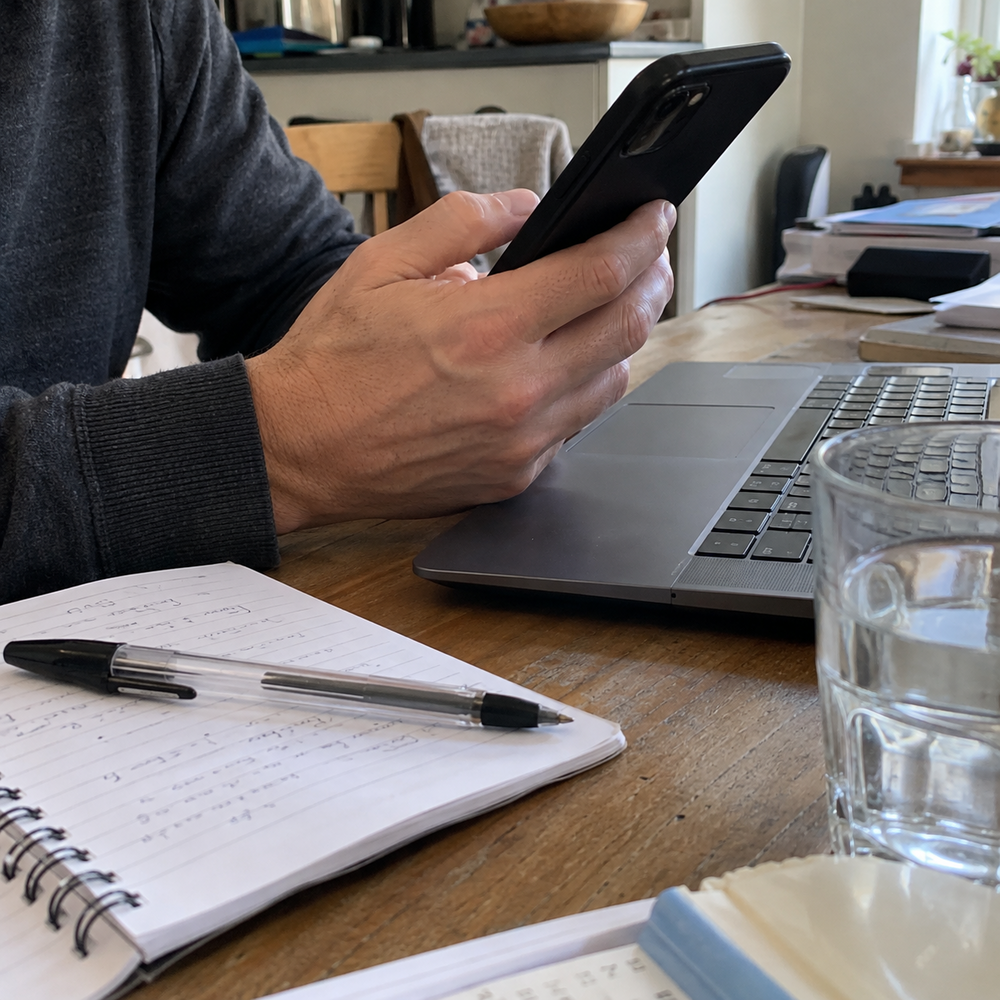Adult holding a smartphone beside a laptop and notebook at a kitchen table.