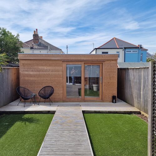 A sleek and modern garden room designed and installed by Forever Timber, supplied by Lugarde. It features a natural wood finish with a flat roof, large sliding glass doors, and a clean, minimalist design. The space is complemented by a decked area at the front, ideal for outdoor seating, and a pathway leading through a well-maintained artificial lawn. Perfect for a home office, studio, or relaxing retreat.