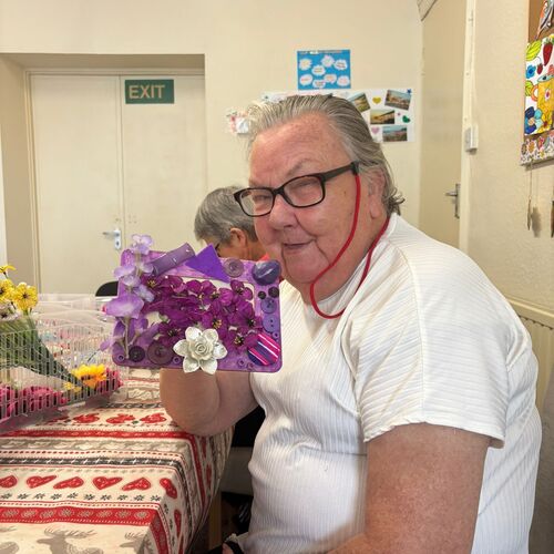 Elderly lady smiling at the table, holding her frame that she decorated at her arts and crafts session at Your Care's Dementia Day Centre, Bristol