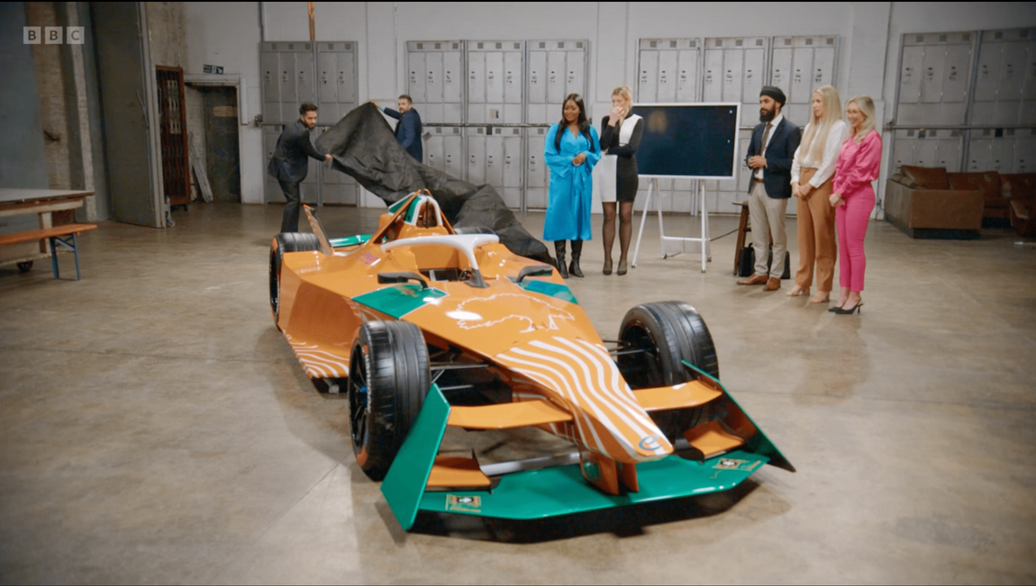 A group of people standing around a race car on the apprentice